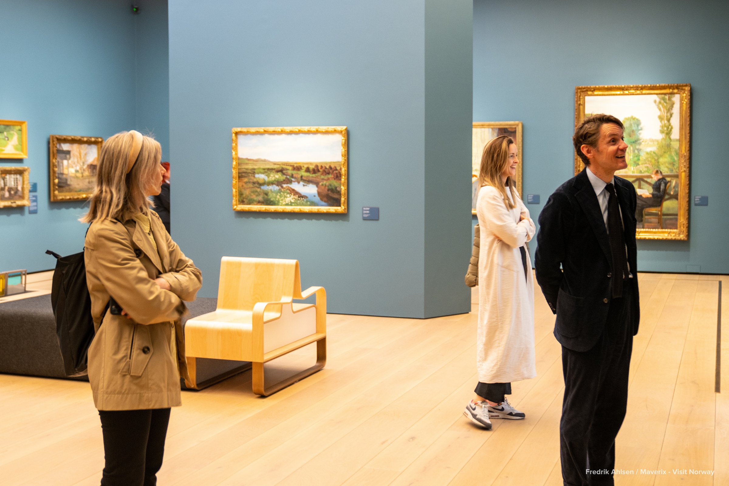 People admire framed landscape paintings in a brightly lit room with blue walls.