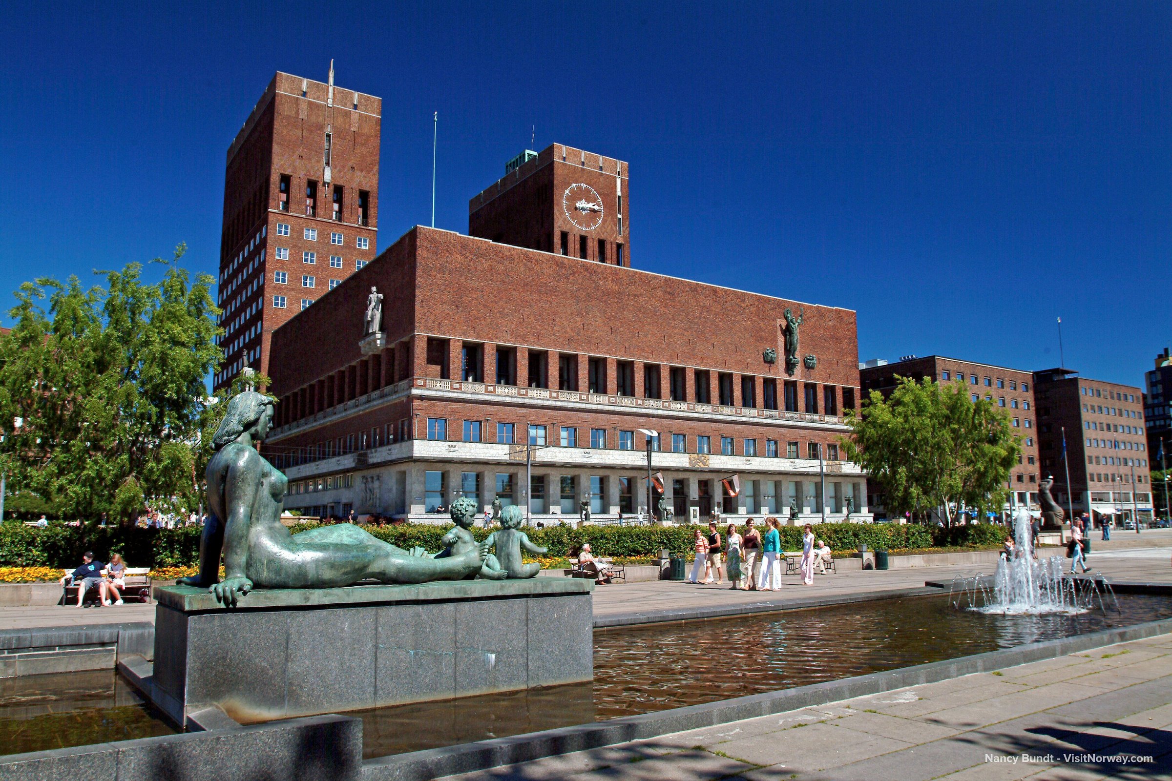 Red brick Oslo City Hall with statues and fountains in the foreground under a clear blue sky.