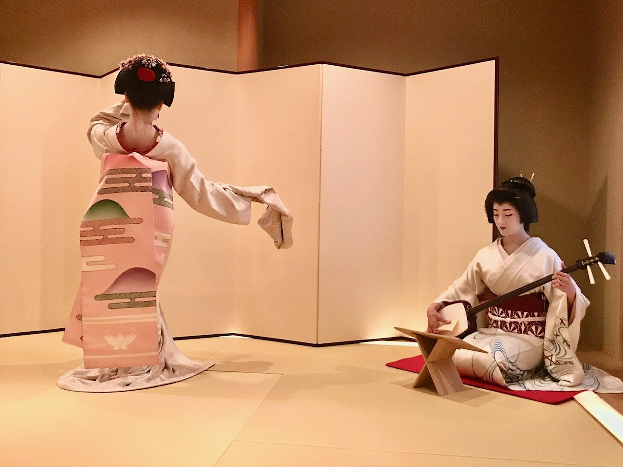 A geisha in a white kimono performs a traditional dance while another sits nearby playing a shamisen, both set against a minimalist folding screen in a tatami-mat room.